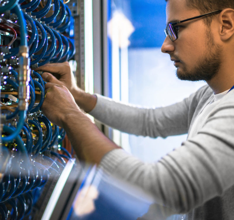Side view portrait of young man wearing glasses connecting cables in server cabinet while working with supercomputer in data center